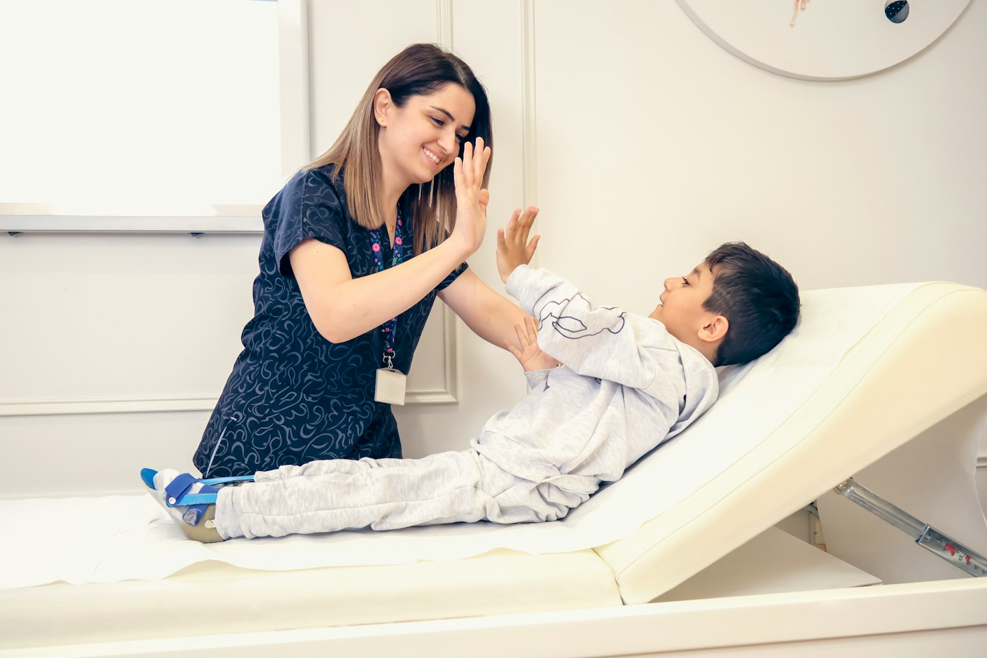 Eine Frau gibt einem Jungen mit einer Beinorthese auf einem Untersuchungstisch einen High-Five.